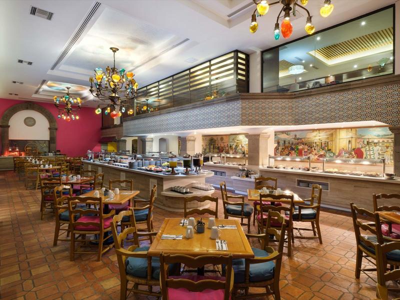 Grand restaurant avec tables en bois, chaises colorées et un grand buffet sous une galerie.