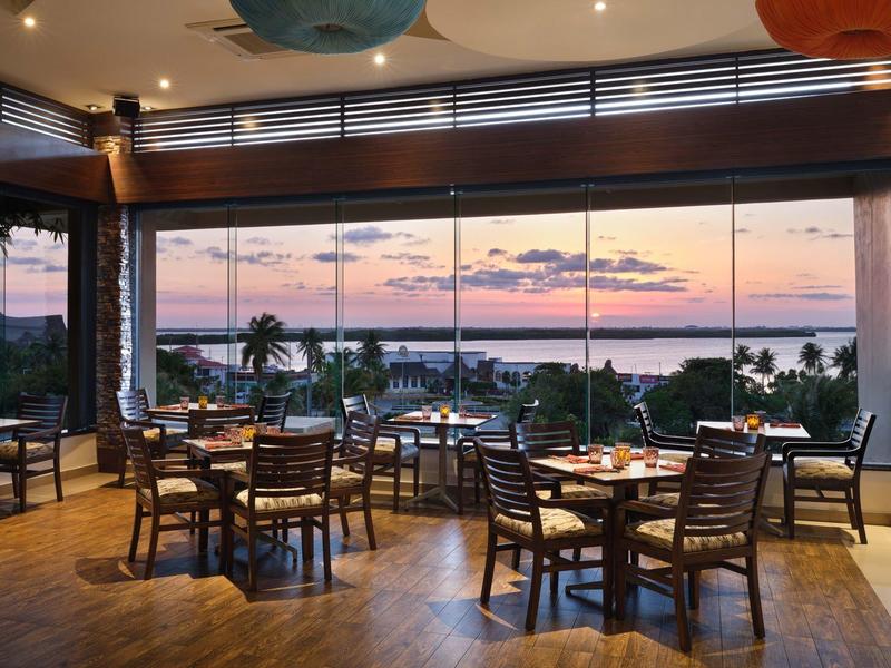 Restaurant ouvert avec des chaises et tables en bois avec vue sur un coucher de soleil tropical et des palmiers.