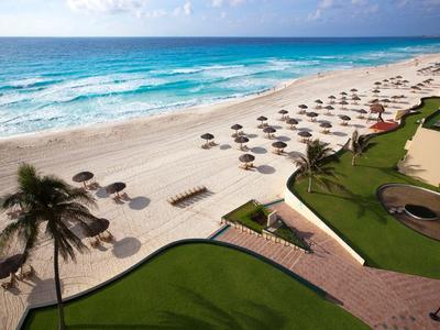 Breiter Sandstrand mit Sonnenschirmen, blauem Meer und Palmen hinter gepflegtem Rasen.