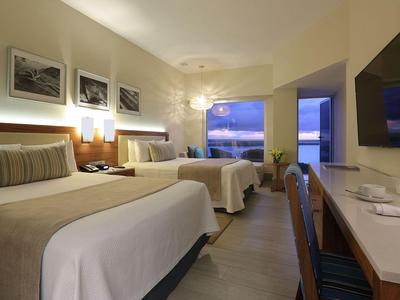 Habitación de hotel moderna con dos camas, escritorio, TV y vistas al mar al atardecer.