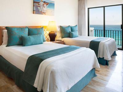 Habitación de hotel luminosa con dos camas dobles, cojines azules y vistas al mar por ventanales grandes