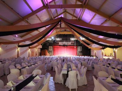 Grote evenementenhal met gedecoreerde tafels en stoelen voor een rood podium onder paarse verlichting.