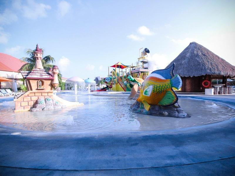 Espace piscine adapté aux enfants avec fontaine poisson colorée et aire de jeux aquatique en arrière-plan.