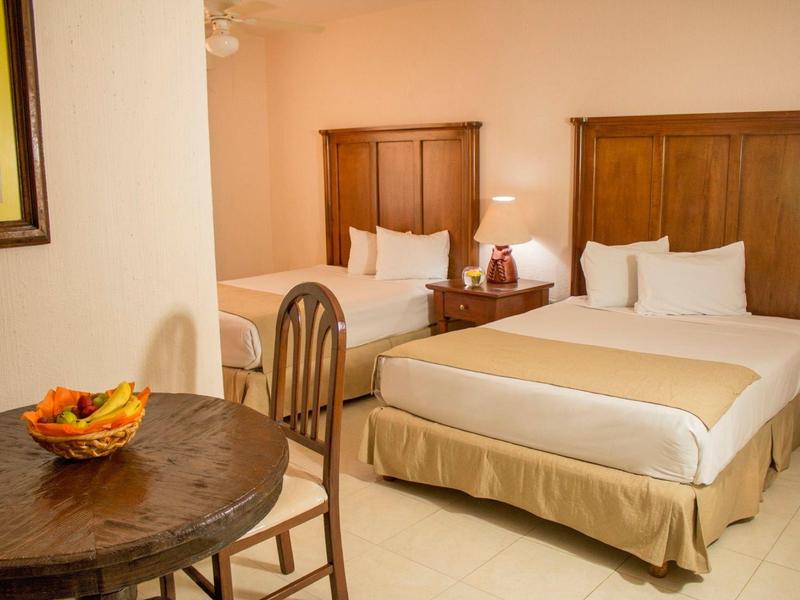 Chambre d'hôtel lumineuse avec deux lits doubles, meubles en bois et une table avec un bol de fruits.