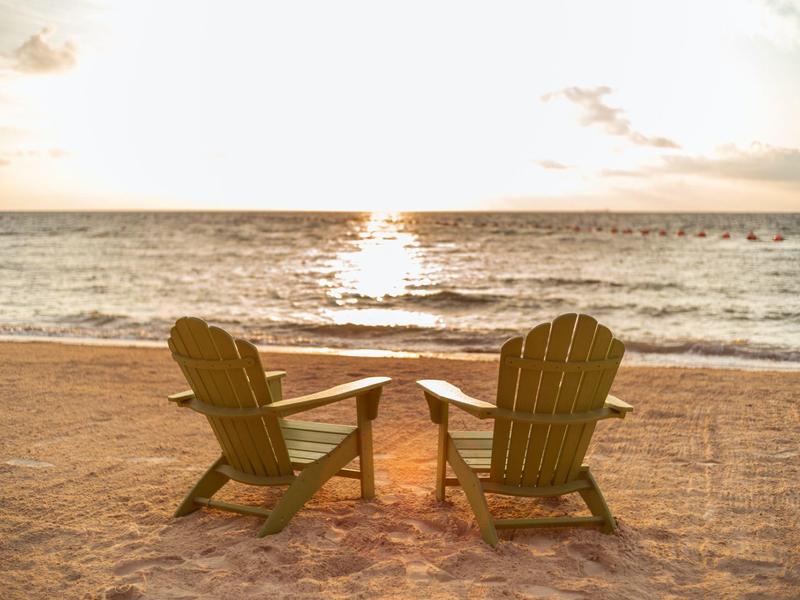 Due sedie di legno con un piccolo tavolo sulla spiaggia con mare calmo e tramonto.