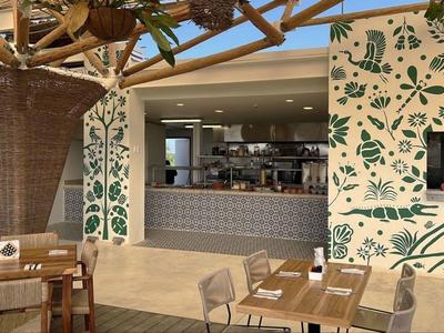 Moderner Cafébereich mit Holztischen, geflochtener Säule und Wand mit grünem Blättermuster.