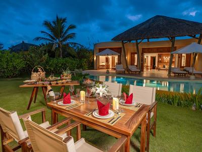 Een gedekte eettafel met rode servetten in de tuin voor een verlicht huis en zwembad bij schemering.