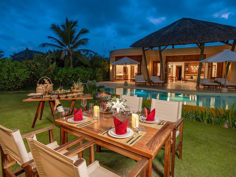 Een gedekte eettafel met rode servetten in de tuin voor een verlicht huis en zwembad bij schemering.