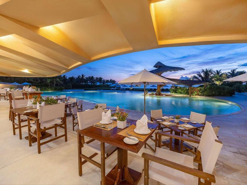 Elegant restaurant met houten tafels en uitzicht op een verlicht zwembad bij zonsondergang.