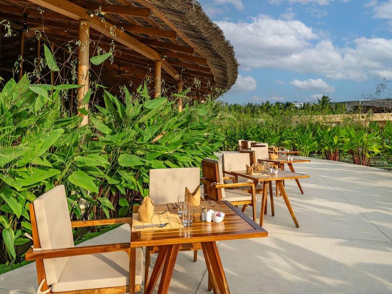 Zonnig terras met houten tafels en stoelen naast groene planten onder blauwe lucht.