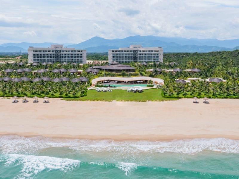 Groot resorthotel met zwembad aan een breed zandstrand en groene achtergrond.