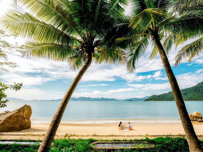 Plaża z palmami, piaskiem i spokojnym morzem pod częściowo zachmurzonym niebem.