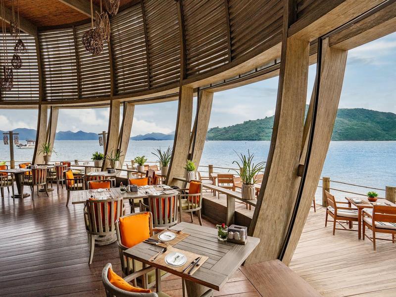 Otwarta restauracja z widokiem na morze, drewniana podłoga, drewniane meble i pomarańczowe akcenty.