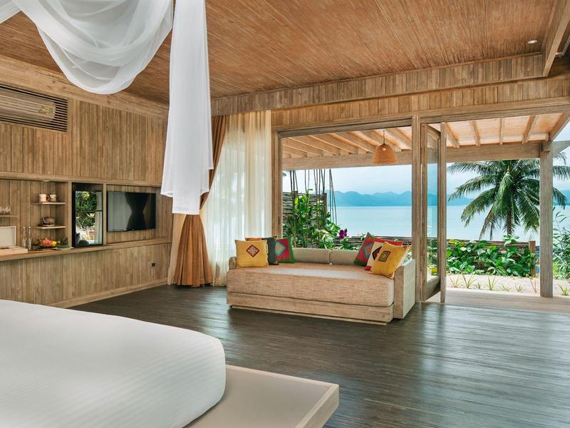 Otwarty pokój hotelowy z drewnianymi ścianami, sofą, łóżkiem i widokiem na morze z palmami.
