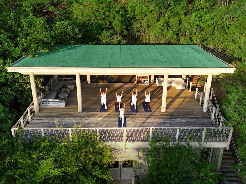 Drewniany zadaszony taras otoczony zielenią z osobami praktykującymi jogę.