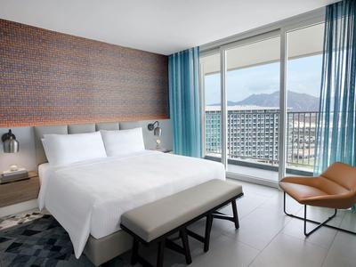 Helles Schlafzimmer mit großem Bett, brauner Lederstuhl, blauen Vorhängen und Panoramafenster.