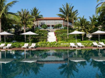 Großer Pool mit acht Liegestühlen und Sonnenschirmen vor tropischem Garten und Haus mit rotem Dach.