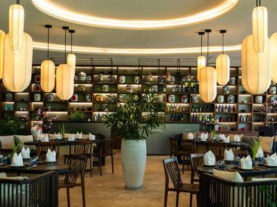 Elegantes Restaurant mit runden Tischen, Holzmöbeln, großer Pflanzenvase und warmen Deckenlampen.