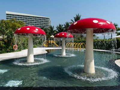 Schwimmbecken mit großen rot-weißen Pilz-Skulpturen und grüner Bepflanzung im Hintergrund.