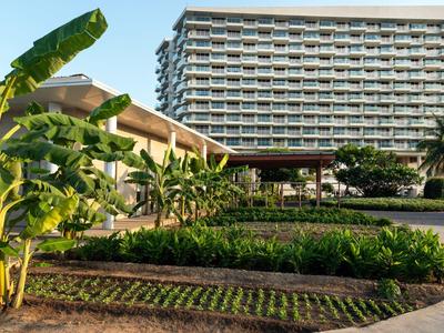 Hotelgebäude mit angrenzendem Garten und tropischen Pflanzen unter blauem Himmel.