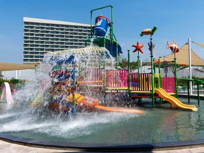 Wasserspielplatz mit Rutsche und Wasserfall vor einem hohen Hotelgebäude bei klarem Himmel.
