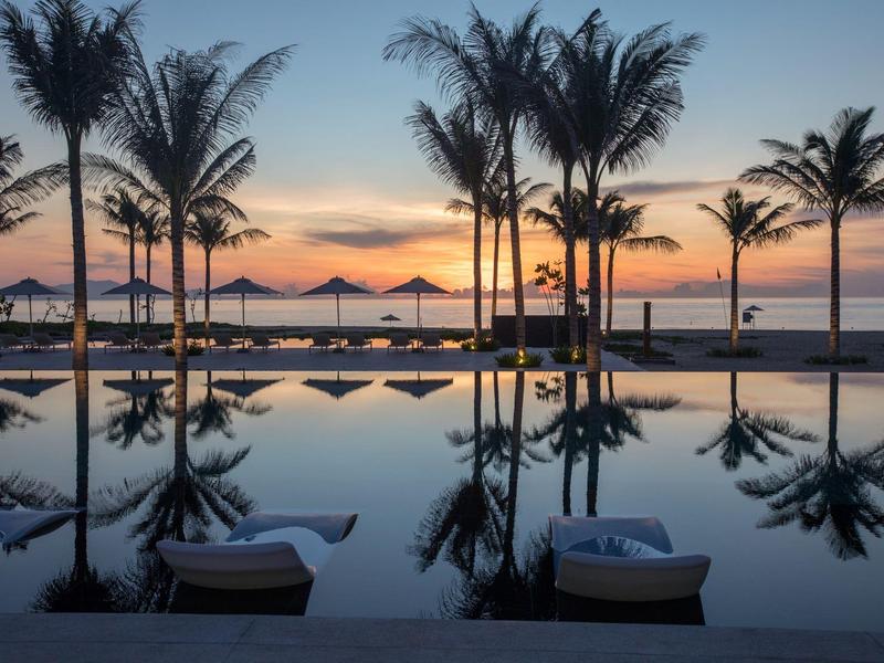 Palmen und Liegen spiegeln sich im Wasser eines Pools bei Sonnenuntergang am Strand.