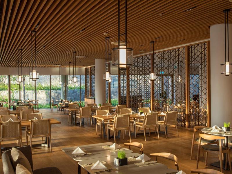 Großes Restaurant mit Holzmöbeln, warmem Licht und bodentiefen Fenstern. Ruhige Atmosphäre.