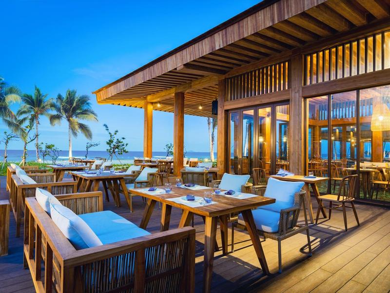 Offene Terrasse mit Holzmöbeln, blauen Kissen, Palmen und Meerblick bei klarem Himmel.