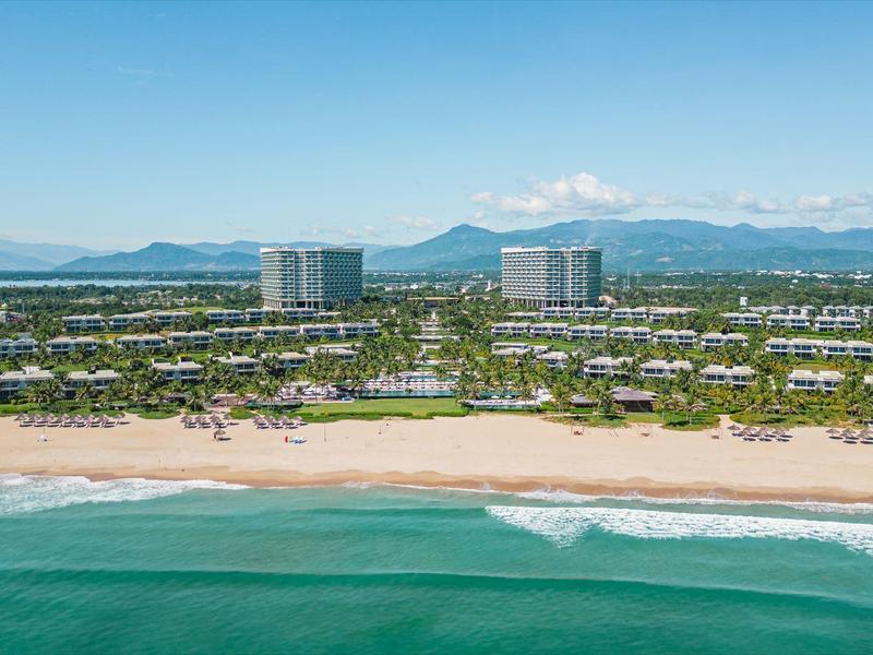 Strand mit grünem Wasser, goldfarbenem Sand und dahinter Häuser, Hochhäuser, Berge im Hintergrund.