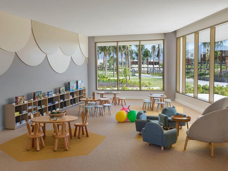 Heller Kinderraum mit Holzmöbeln, bunten Bällen, gemütlichen Sesseln und großen Fenstern mit Blick auf Palmen.