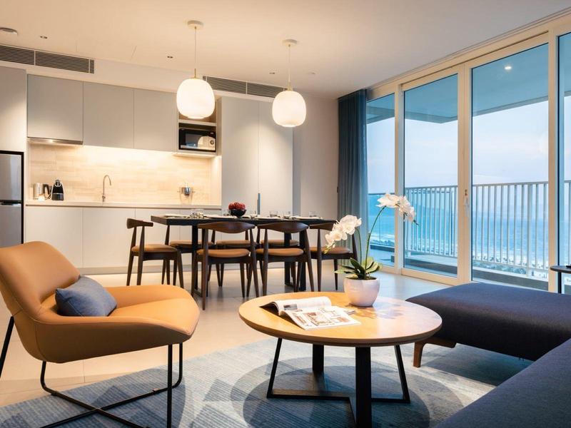 Moderner Wohnraum mit braunem Sessel, rundem Holztisch, Essbereich und großem Glasfenster zum Balkon.