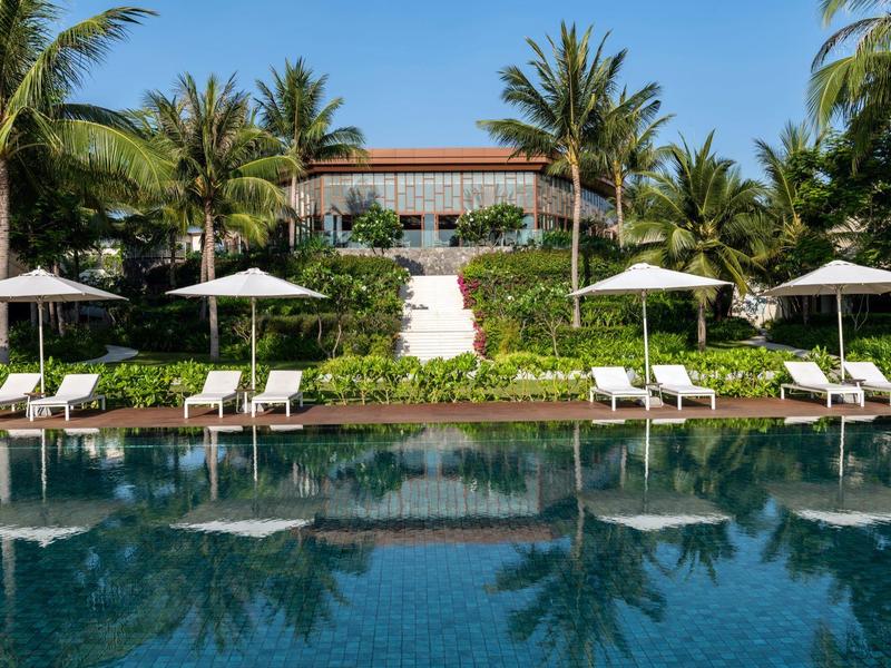 Großer Pool mit acht Liegestühlen und Sonnenschirmen vor tropischem Garten und Haus mit rotem Dach.