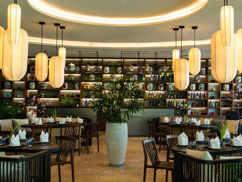 Elegantes Restaurant mit runden Tischen, Holzmöbeln, großer Pflanzenvase und warmen Deckenlampen.