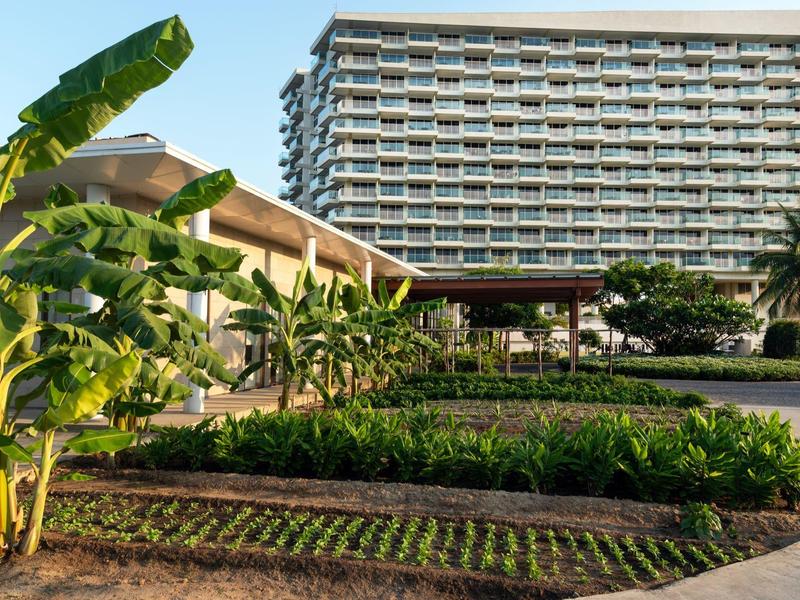 Hotelgebäude mit angrenzendem Garten und tropischen Pflanzen unter blauem Himmel.
