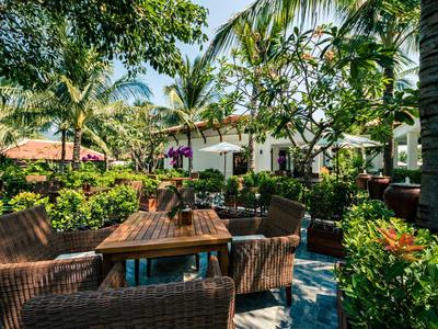 Gemütliche Außensitzgruppe aus Korbmaterial um Holz­tisch, umgeben von grünen Palmen und blühenden Pflanzen.