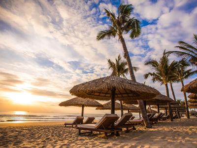 Strand mit Sonnenliegen und Strohdächern bei Sonnenuntergang, Palmen im Hintergrund.