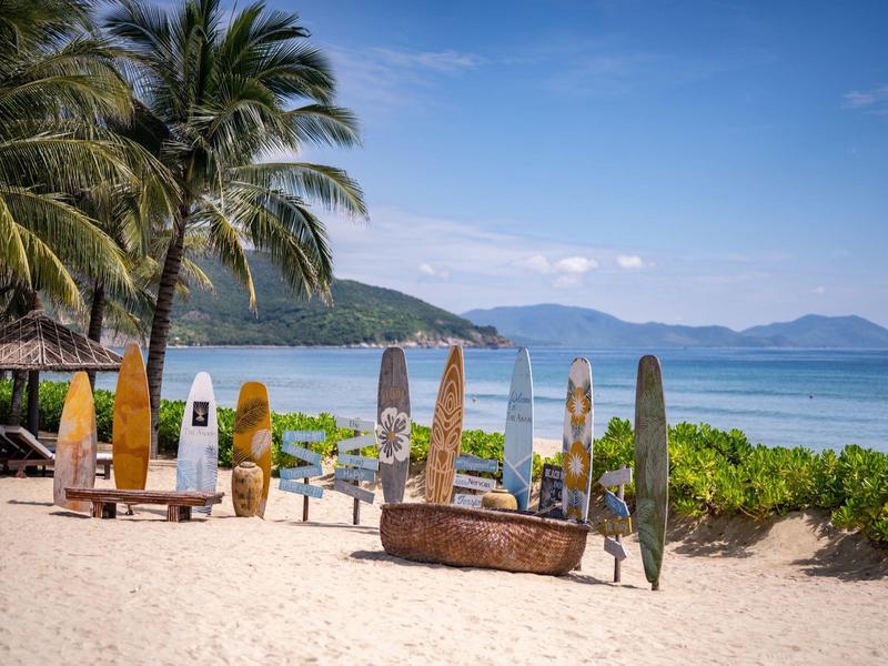 Strand mit Palmen, Sand, mehreren Surfbrettern und ruhigem Meer mit Bergen im Hintergrund.