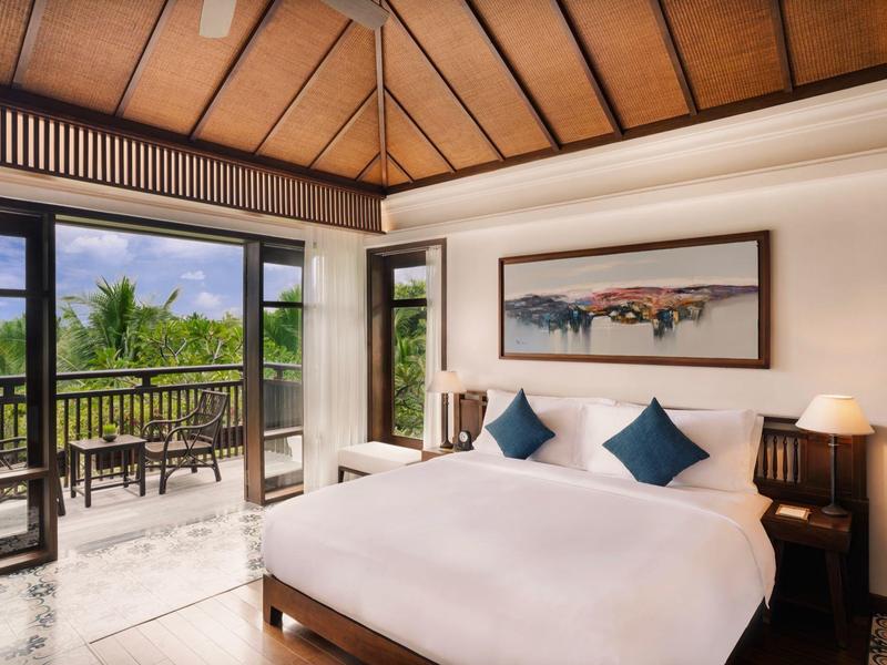 Helles Hotelzimmer mit großem Bett, blauen Kissen und Balkon mit Blick ins Grüne.