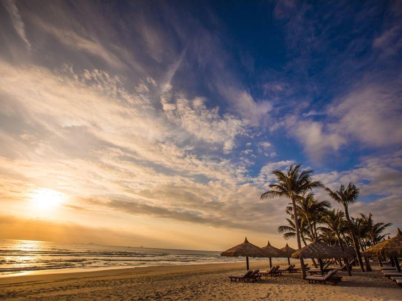 Strand mit Palmen und Sonnenschirmen bei Sonnenuntergang unter einem bewölkten Himmel.