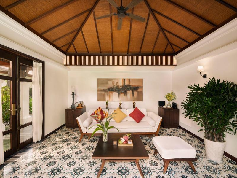 Helles Wohnzimmer mit weißem Sofa, bunten Kissen, großem Pflanzenkübel und Holztisch auf gemustertem Boden.