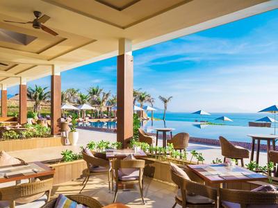 Offener Hotelrestaurantbereich mit Blick auf den Strand und das Meer bei sonnigem Wetter.