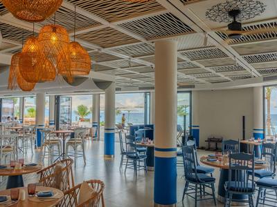 Restaurant d'hôtel lumineux avec mobilier en bois et lampes suspendues en osier, vue sur la mer par les fenêtres.