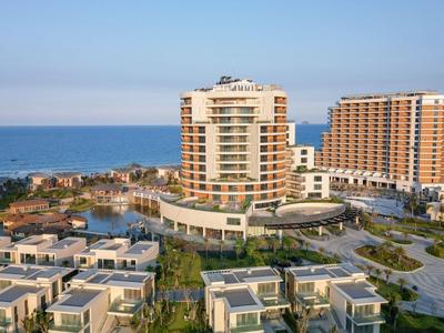 Complexe hôtelier moderne en bord de mer avec piscine et vue sur la mer sous un ciel clair.