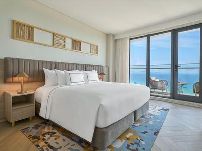 Chambre d'hôtel moderne avec grand lit, table de nuit, tapis et vue sur la mer par des fenêtres du sol au plafond.
