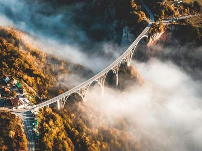 Luftaufnahme einer Brücke durch nebligen Wald mit herbstlichen Farben und einem nahegelegenen Dorf.