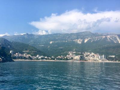 Blick auf ein Küstenhotel vor bewaldeten Bergen und klarem Himmel über ruhigem Wasser.