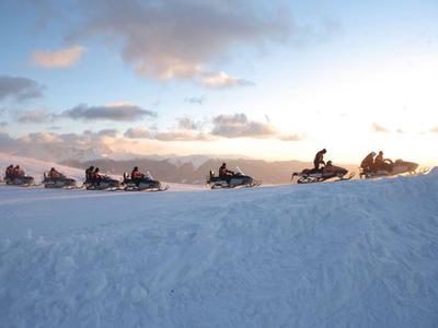 Mehrere Schneemobile fahren bei Sonnenuntergang auf einer verschneiten Anhöhe.