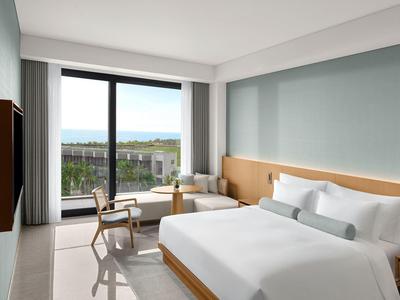 Habitación de hotel luminosa con cama grande, área de estar y balcón con vista a áreas verdes.