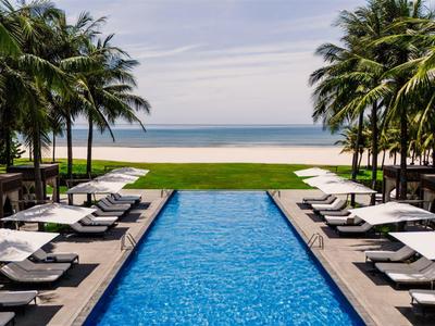 Vista de una piscina con tumbonas y palmeras, con la playa y el mar al fondo.