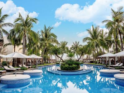 Großer blauer Pool umgeben von Palmen, weißen Sonnenbetten und Sonnenschirmen unter blauem Himmel.